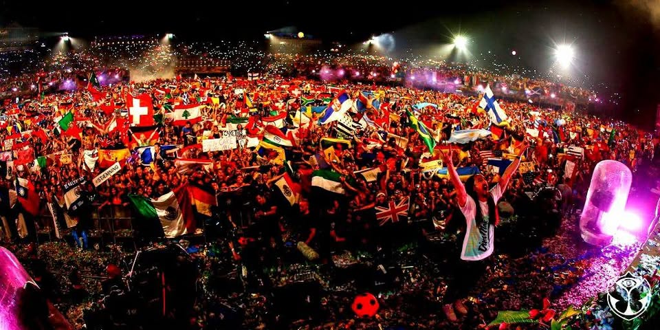 “Wide night-time shot of a packed outdoor concert crowd waving Lebanese and international flags under bright stage lights.”  “صورة بانورامية لحشد جماهيري كثيف في حفل موسيقي ليلي بالهواء الطلق يلوّح بالأعلام اللبنانية والدولية تحت أضواء المسرح.